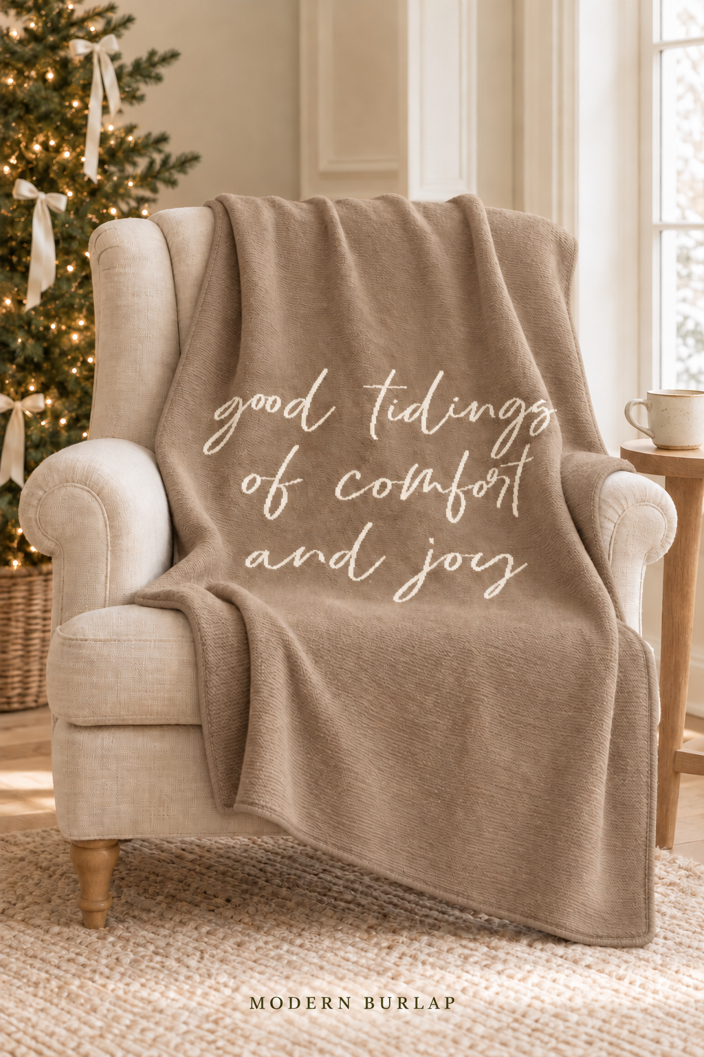 Good Tidings of Comfort and Joy throw in Taupe, draped over an ivory armchair beside a Christmas tree. Matching taupe border. Lifestyle styling.