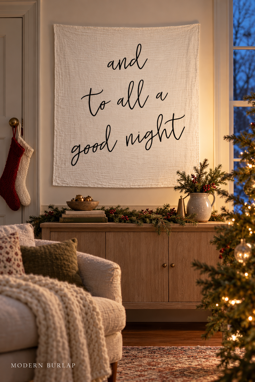 And to All a Good Night white muslin swaddle above an oak cabinet beside Christmas stockings and a blue-hour window. Modern Burlap wordmark at the bottom.
