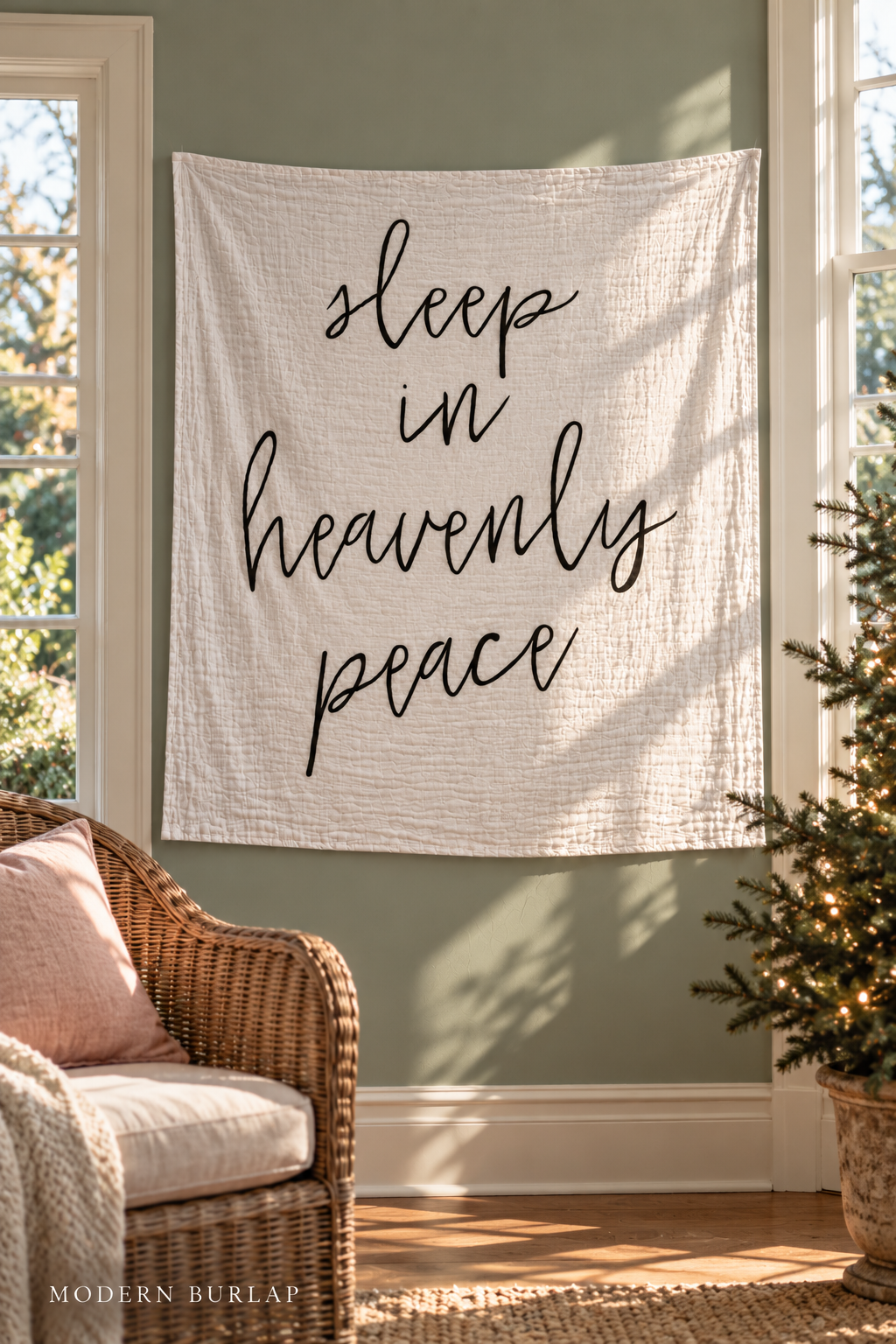 Sleep in Heavenly Peace white muslin swaddle on a green sunroom wall between windows beside a wicker chair and fir tree. Modern Burlap wordmark at the bottom.