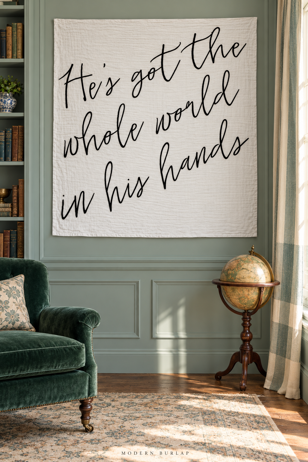 A square white He’s Got the Whole World muslin swaddle hangs in a celadon library beside an emerald chair and small antique globe.