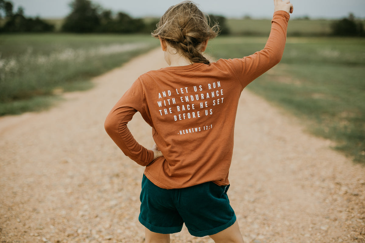 Child wearing the Hebrews 12:1 long sleeve tee with the printed back visible on a country road.
