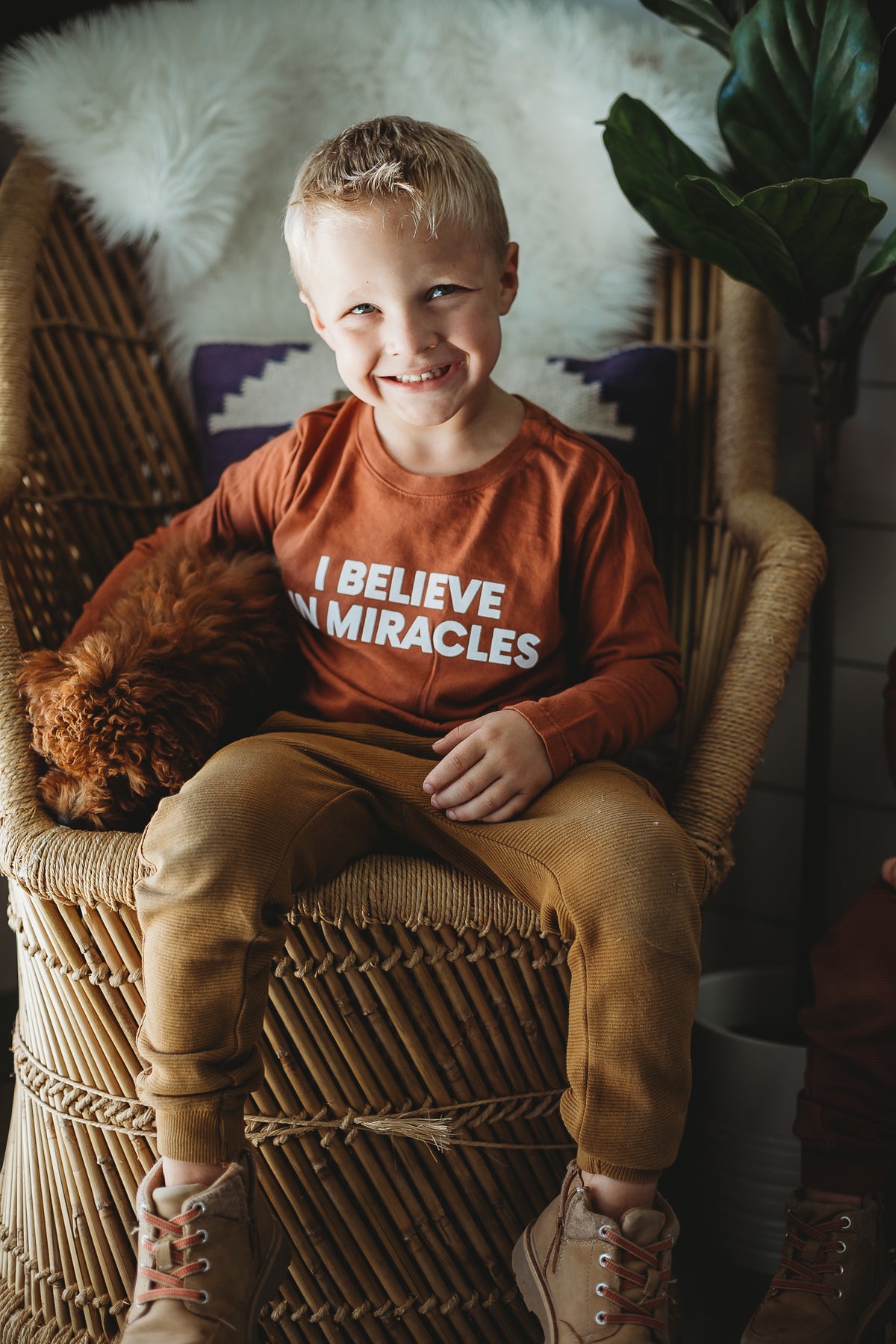 Child seated in a woven chair wearing the Sunburn I Believe in Miracles long sleeve tee.