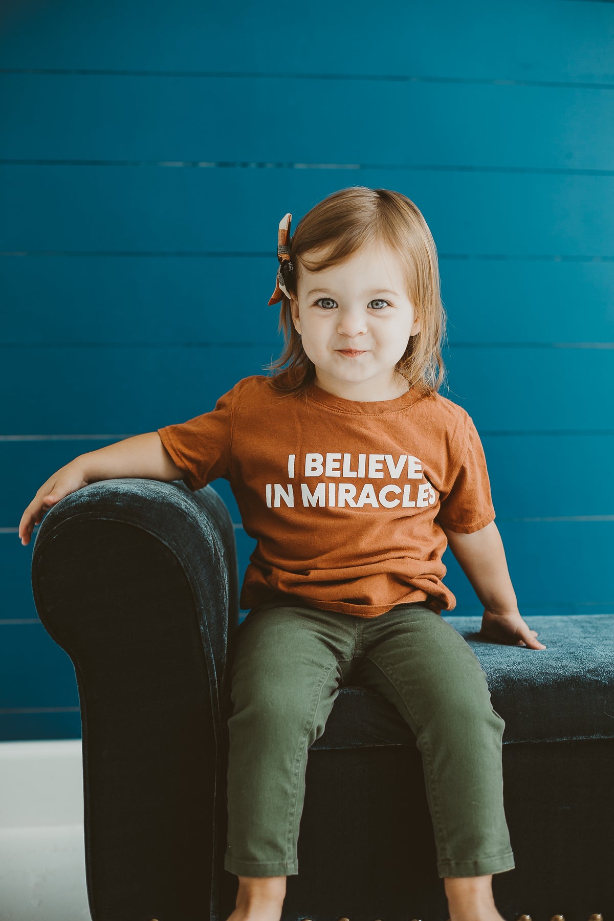 Child seated in a dark chair wearing the Sunburn I Believe in Miracles short sleeve tee.