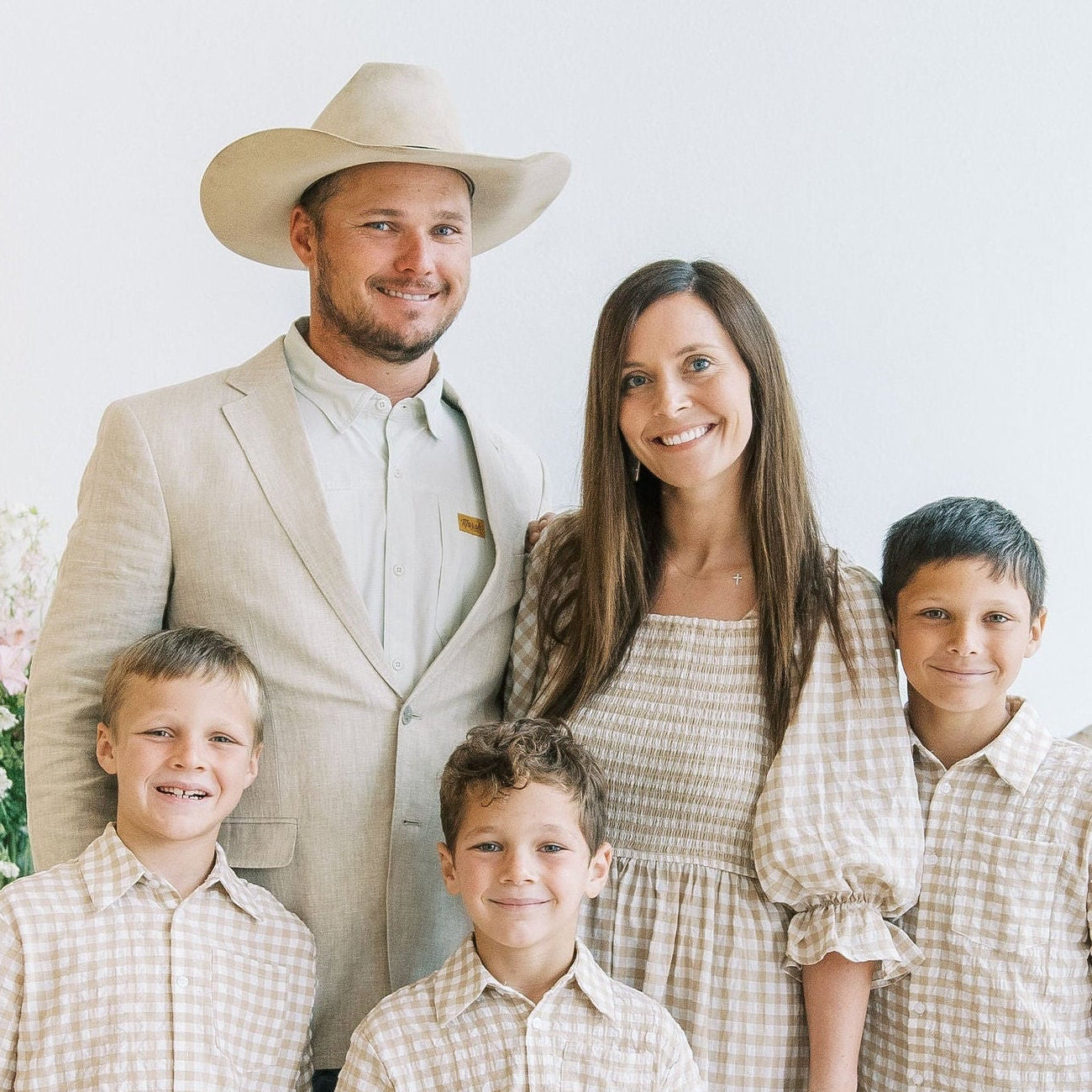 A family of five posing together in matching beige and white gingham clothing, with the father wearing a light beige suit and cowboy hat, the mother in a matching tiered dress, and the three boys in matching short-sleeve gingham shirts and jeans.