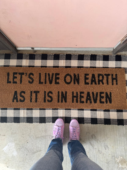 Natural coir doormat reading Let's live on earth as it is in heaven, layered over a checked rug at a pink door.