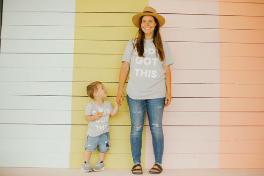 Woman and child standing together in gray God's Got This tees against a pastel wall.