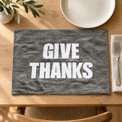 Graphite linen Give Thanks placemat with white lettering on a wood dining table.