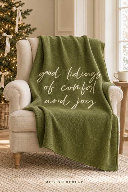 Good Tidings of Comfort and Joy throw in Kaki Green, draped over an ivory armchair beside a Christmas tree. Lifestyle styling.