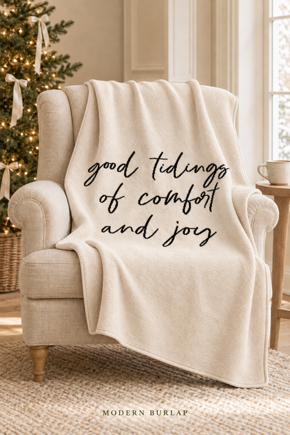 Good Tidings of Comfort and Joy throw in Natural, draped over an ivory armchair beside a Christmas tree. Lifestyle styling.