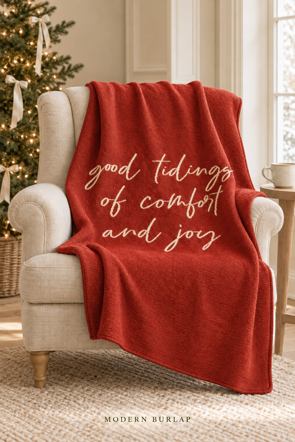 Good Tidings of Comfort and Joy throw in Rust Red, draped over an ivory armchair beside a Christmas tree. Lifestyle styling.