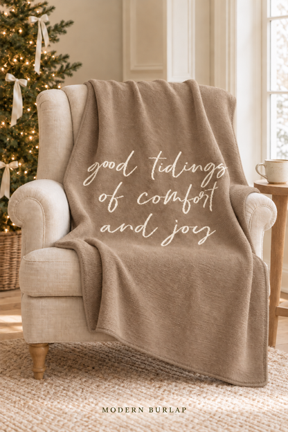 Good Tidings of Comfort and Joy throw in Taupe, draped over an ivory armchair beside a Christmas tree. Matching taupe border. Lifestyle styling.