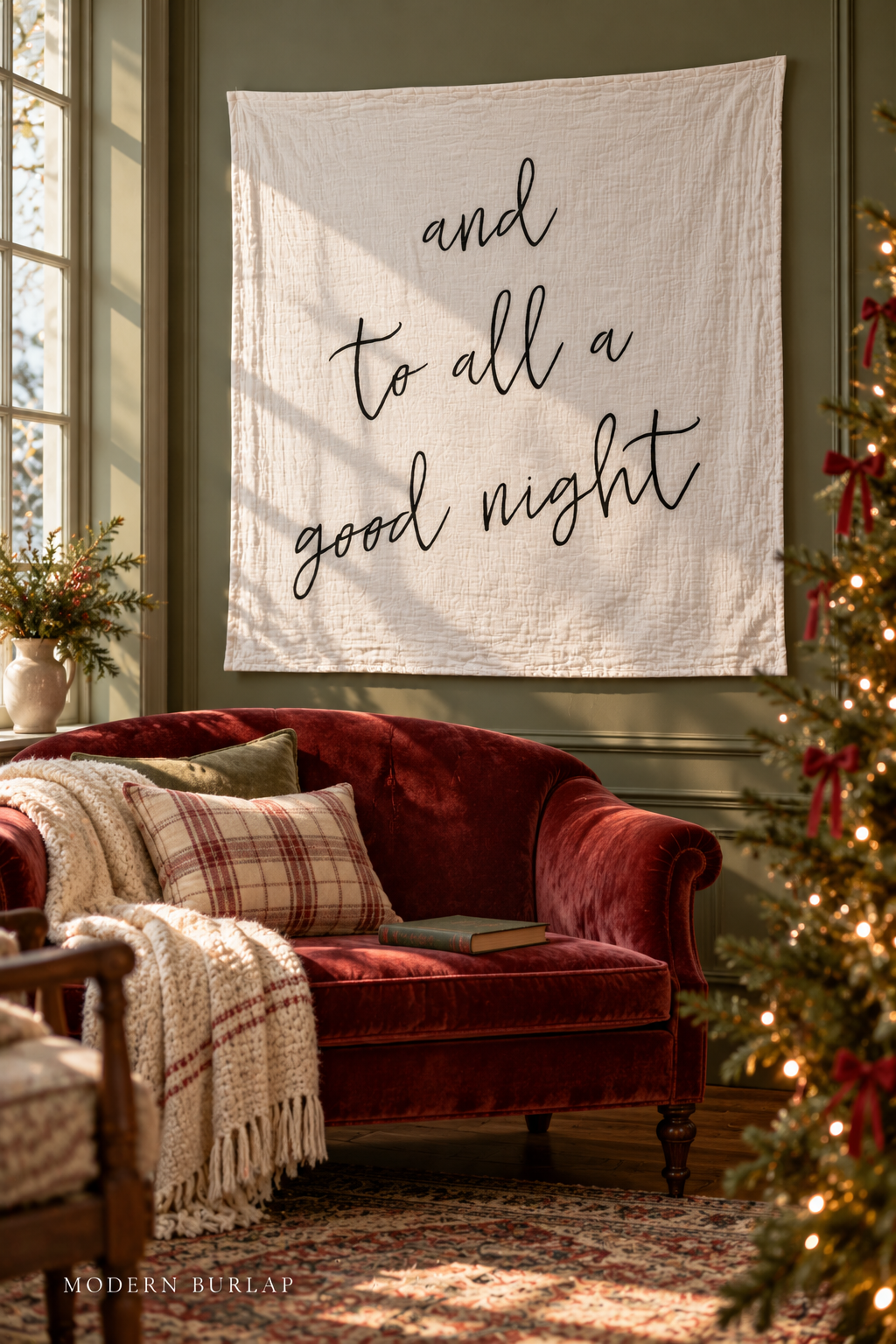 And to All a Good Night white muslin swaddle above a cranberry sofa in a green Christmas reading room. Modern Burlap wordmark at the bottom.