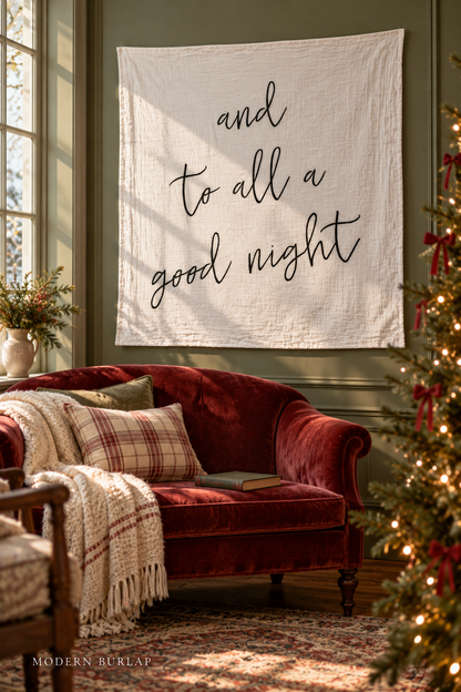 And to All a Good Night white muslin swaddle above a cranberry sofa in a green Christmas reading room. Modern Burlap wordmark at the bottom.