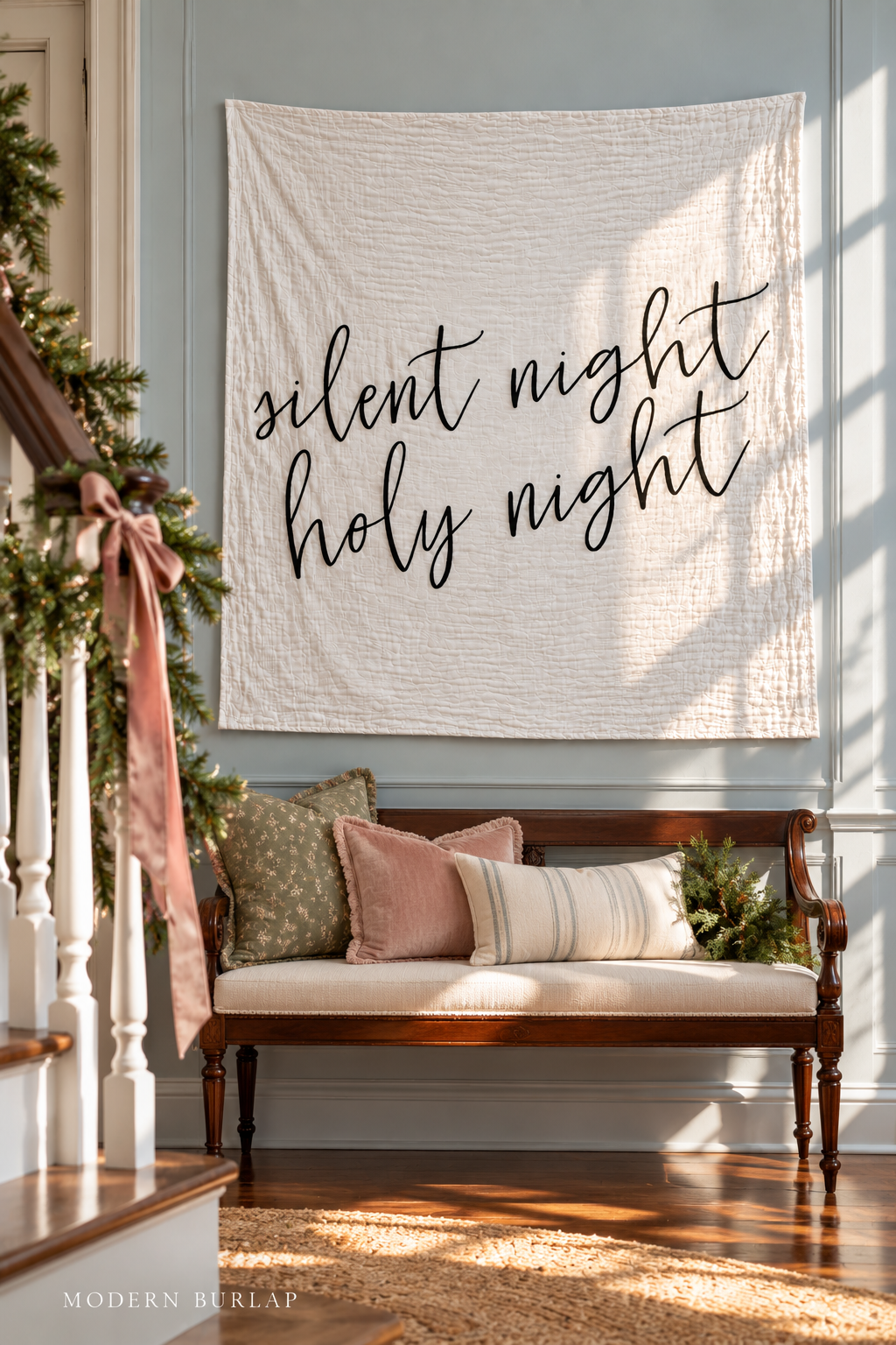Silent Night Holy Night white muslin swaddle above a settee beside a garland-trimmed staircase. Modern Burlap wordmark at the bottom.