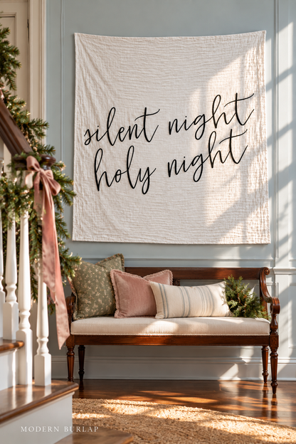 Silent Night Holy Night white muslin swaddle above a settee beside a garland-trimmed staircase. Modern Burlap wordmark at the bottom.