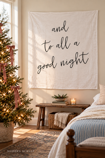 And to All a Good Night white muslin swaddle on a cream wall in a Christmas guest bedroom. Modern Burlap wordmark at the bottom.