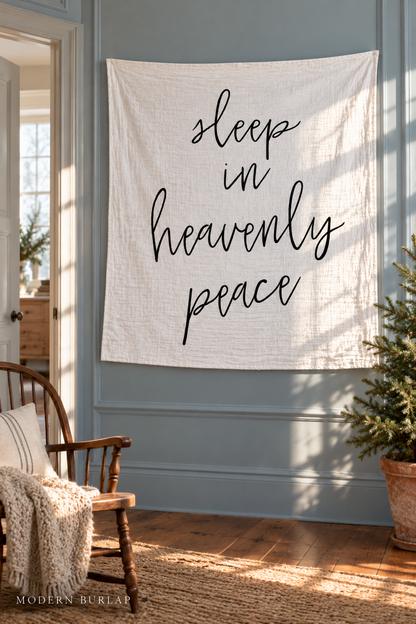 Sleep in Heavenly Peace white muslin swaddle on a blue paneled wall beside a spindle chair and small fir tree. Modern Burlap wordmark at the bottom.