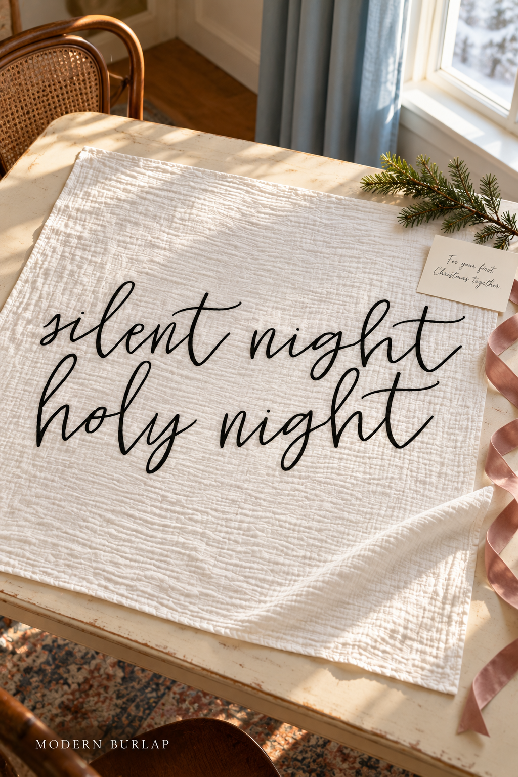 Silent Night Holy Night white muslin swaddle spread on a cream table with a pink ribbon and a handwritten Christmas card. Modern Burlap wordmark at the bottom.