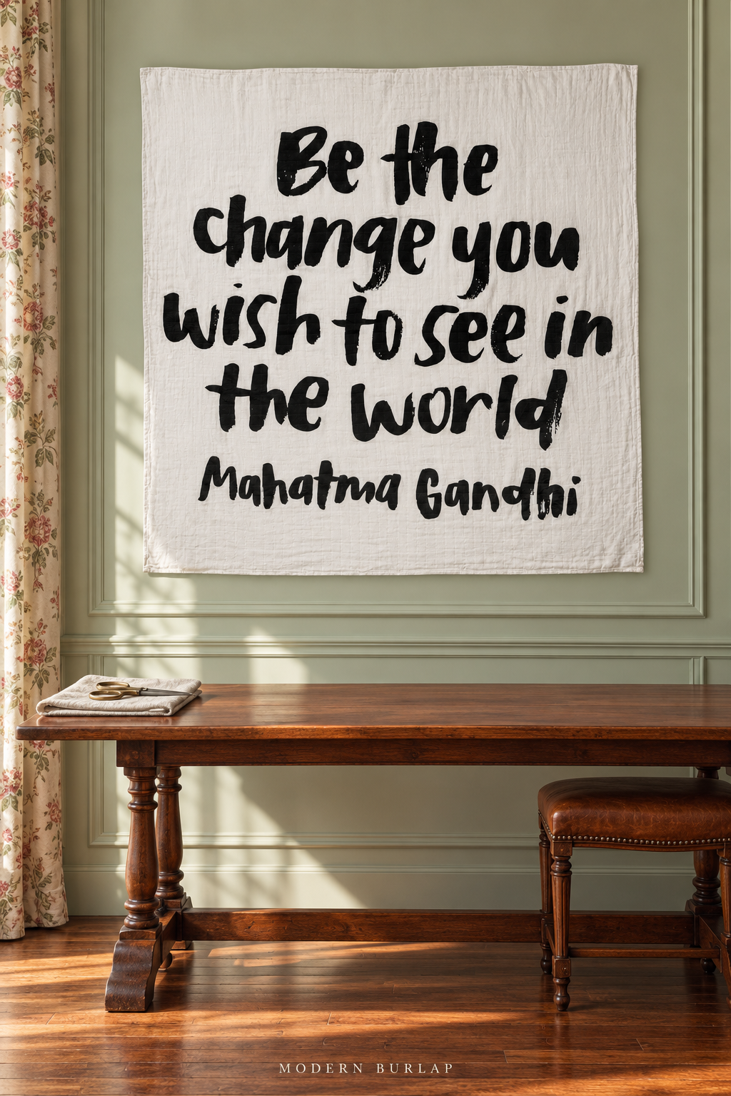 Square white Be the Change muslin wall hanging with black brush lettering and printed Mahatma Gandhi attribution above a mahogany worktable, folded linen and scissors.