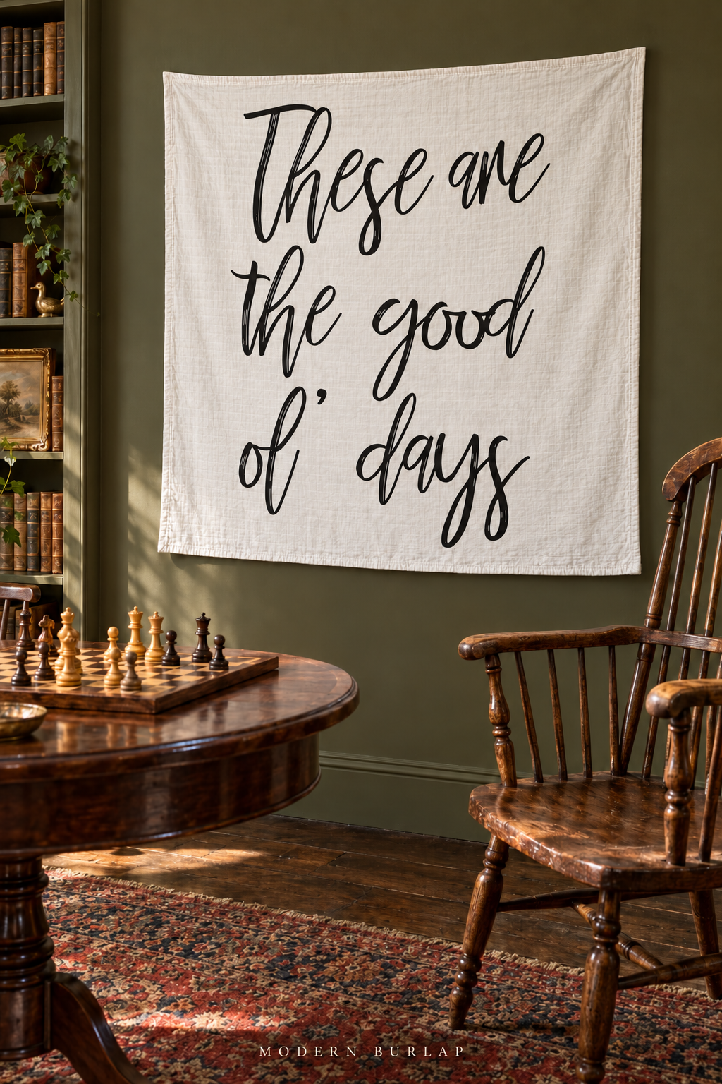 A white These Are the Good Ol’ Days muslin swaddle hangs on an olive wall beside bookshelves, with an antique wooden chess table and spindle chair in the foreground.