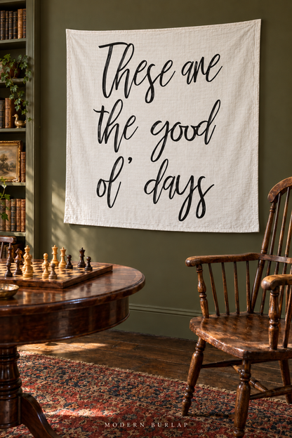 A white These Are the Good Ol’ Days muslin swaddle hangs on an olive wall beside bookshelves, with an antique wooden chess table and spindle chair in the foreground.