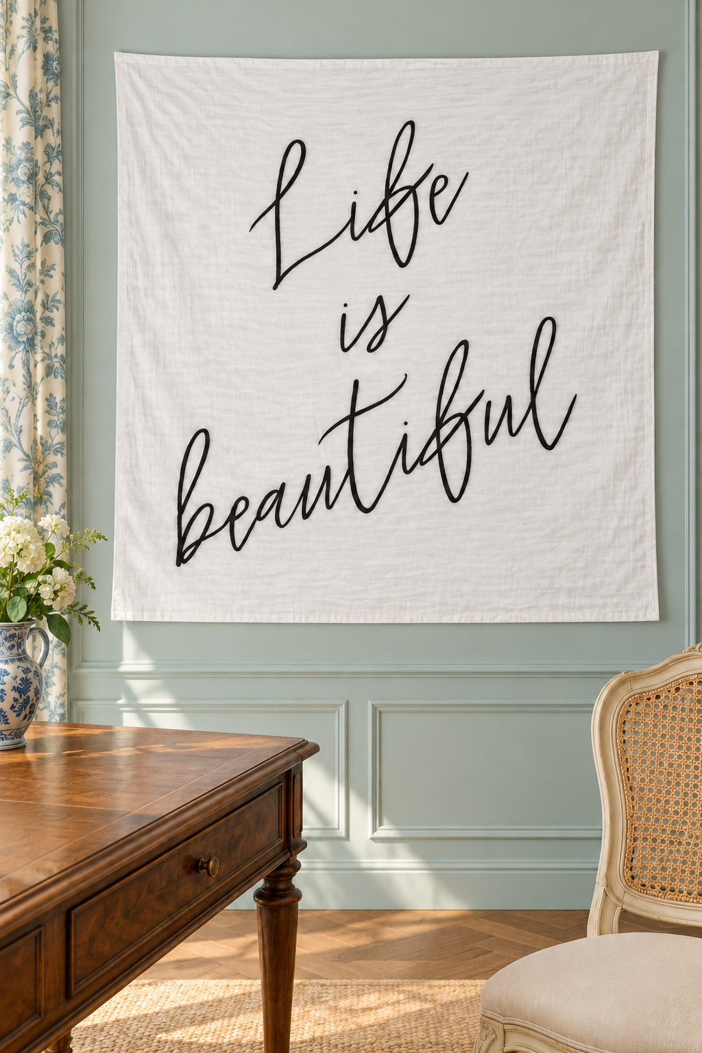 Life Is Beautiful white muslin swaddle displayed on a pale blue-green paneled wall beside a walnut desk and cane chair.