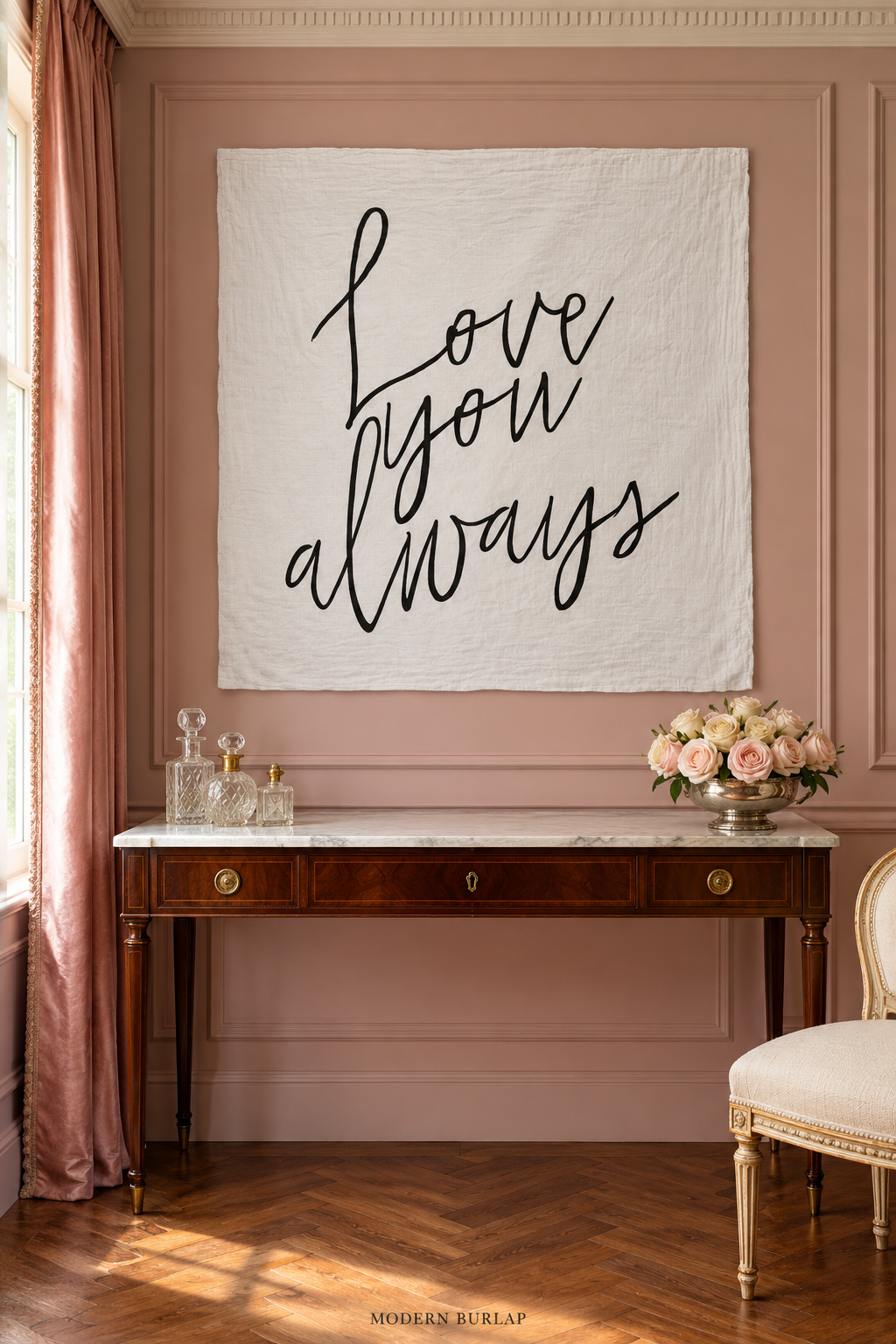 White Love You Always muslin swaddle hanging on a dusty-rose paneled wall above a marble-topped mahogany dressing table, with roses, crystal bottles, and a cream upholstered chair.