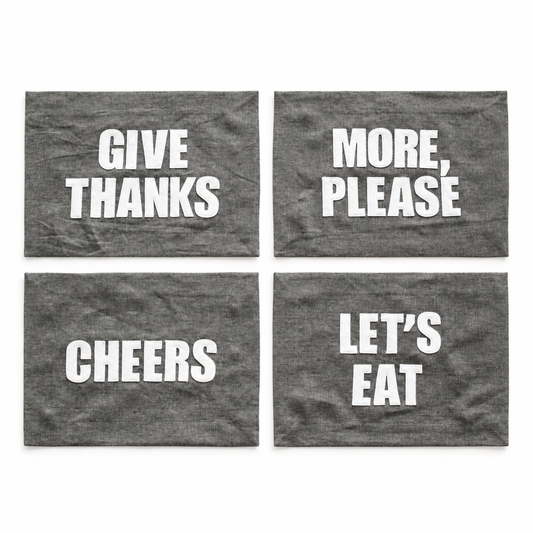 Four graphite linen placemat phrase options: Let's Eat, Cheers, Give Thanks, and More Please. Each sold separately.