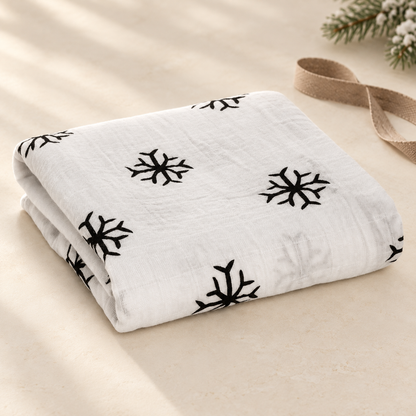 Snowflakes organic cotton muslin swaddle folded on an ivory surface with winter gift styling. Ribbon and greenery are styling props.