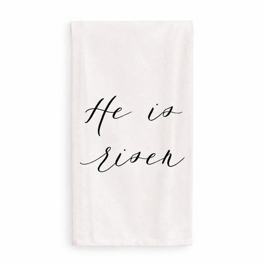 He Is Risen cream cotton tea towel with black script, shown against a white studio background.