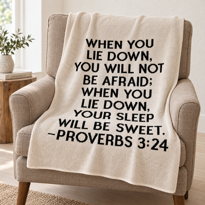 Natural ivory Proverbs 3:24 blanket with black block lettering draped over a taupe linen reading chair.