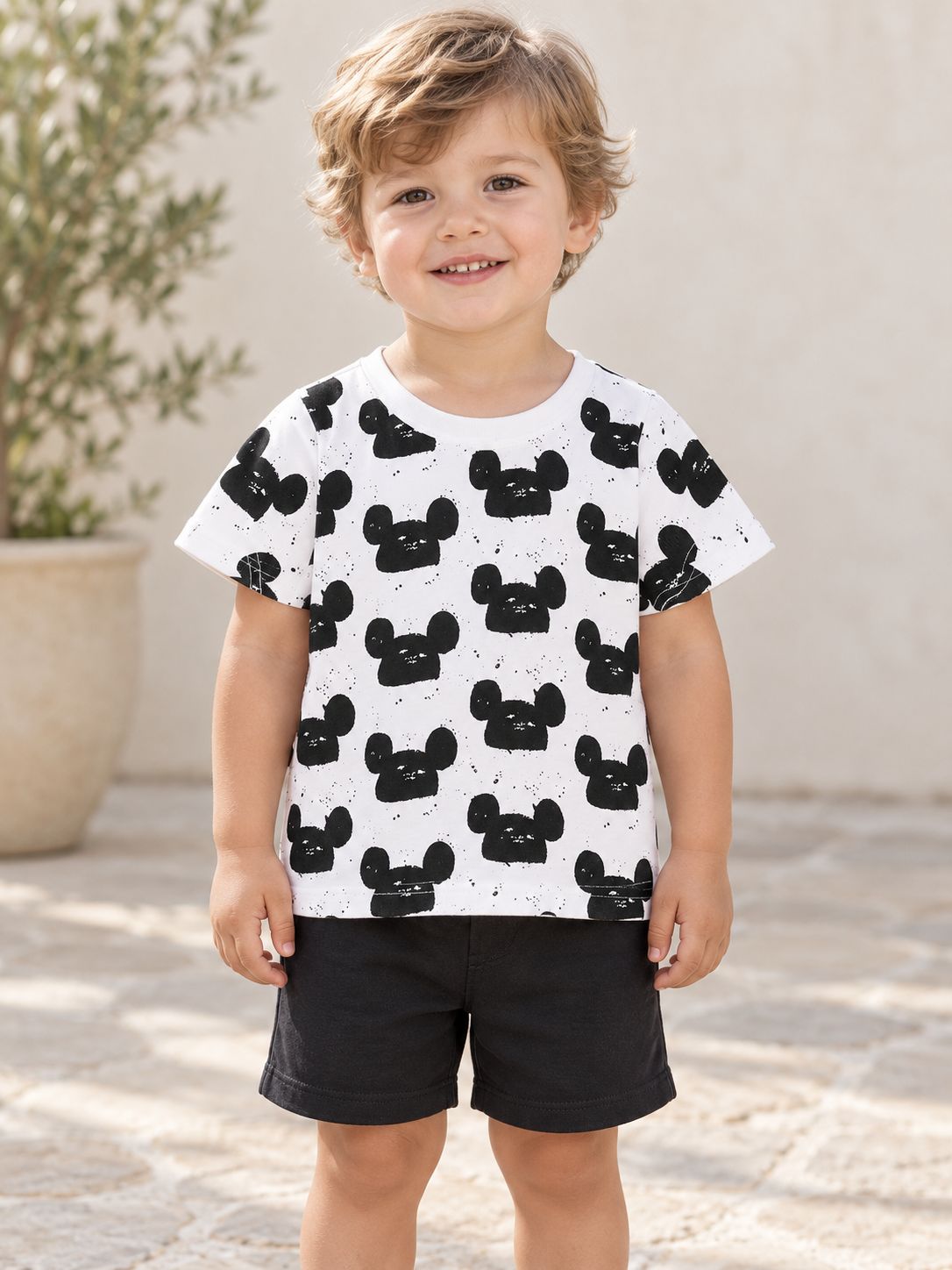 Child wearing a white mouse-head-print tee and black shorts outdoors.