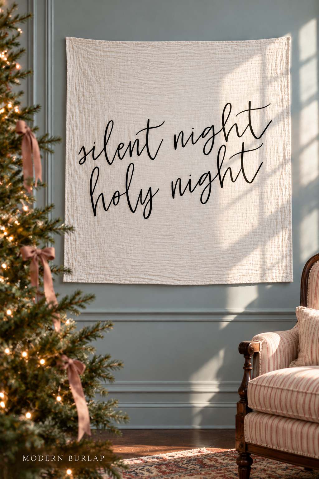 Silent Night Holy Night white muslin swaddle displayed on a blue paneled wall beside a Christmas tree with dusty-rose ribbons and a rose-striped antique chair.
