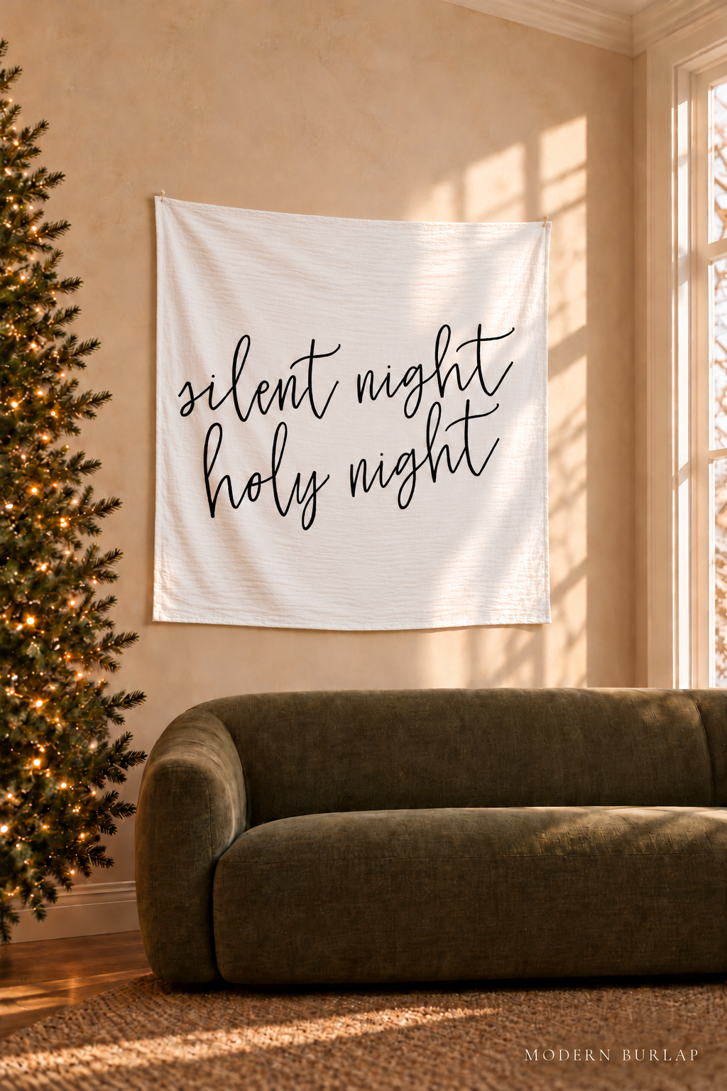 Silent Night Holy Night white muslin swaddle displayed on a warm plaster wall beside a sunlit window, an olive sofa and a Christmas tree.