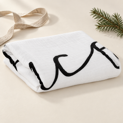 Folded Sleep in Heavenly Peace organic muslin swaddle with ribbon and evergreen styling props; props not included.