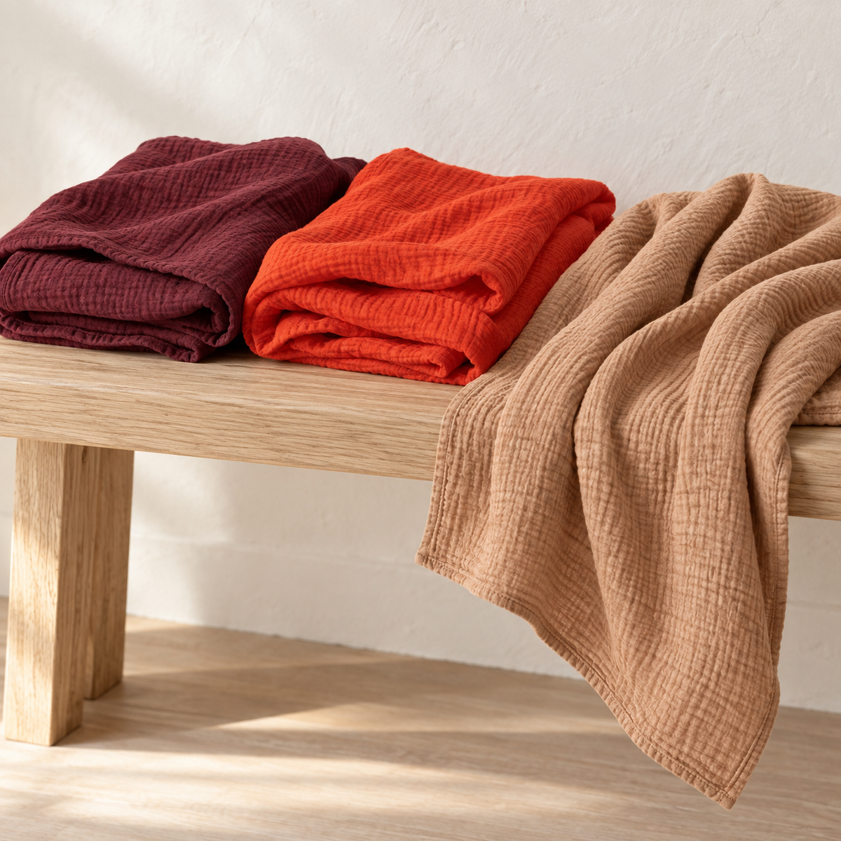 Maroon and Wildfire swaddles folded on an oak bench with Ginger draped to show its lightweight muslin texture.