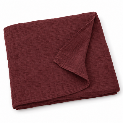 Maroon organic cotton muslin swaddle folded on white, with a turned corner showing the fine weave and hem.