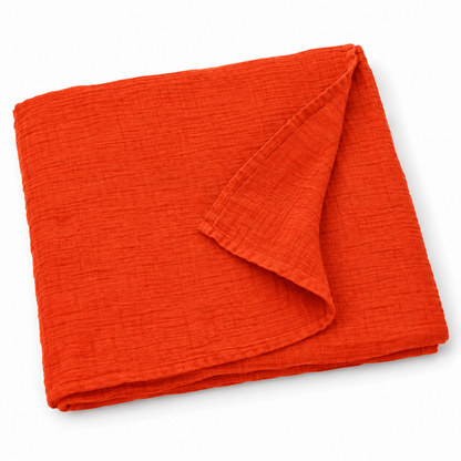 Wildfire orange organic cotton muslin swaddle folded on white, with a turned corner showing the fine weave and hem.
