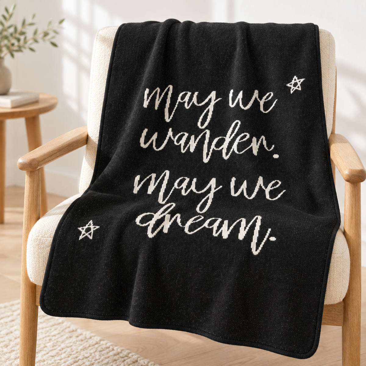 Black May We Wander May We Dream recycled cotton blanket draped over an ivory reading chair with the light script and two stars visible.