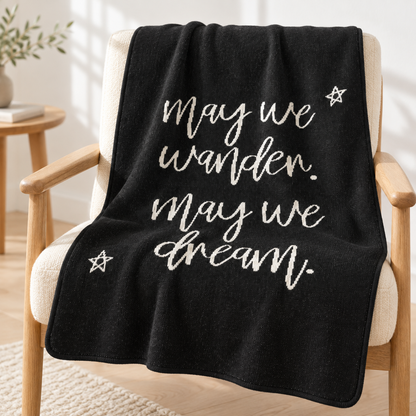 Black May We Wander May We Dream recycled cotton blanket draped over an ivory reading chair with the light script and two stars visible.