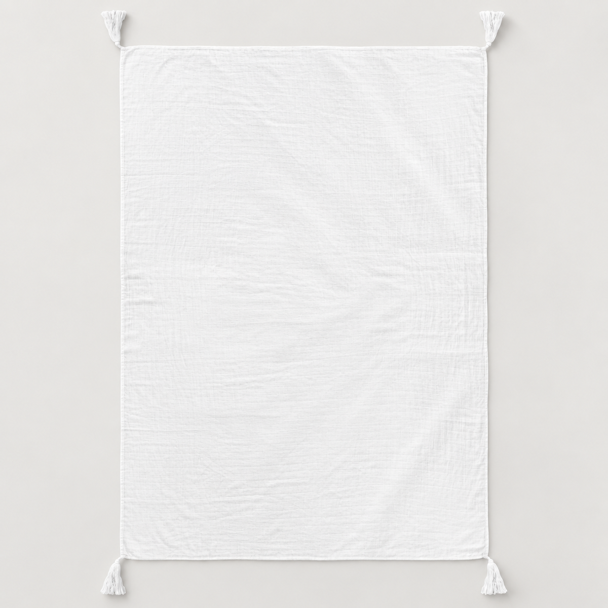White organic cotton muslin throw shown fully open with four corner tassels on a pale gray background.