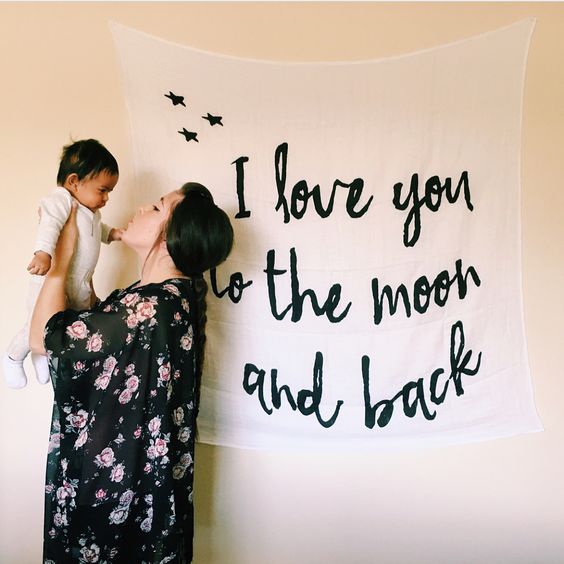 A person holding a baby up to a large swaddle blanket with the text 'I love you to the moon and back' printed on it, along with some star illustrations.