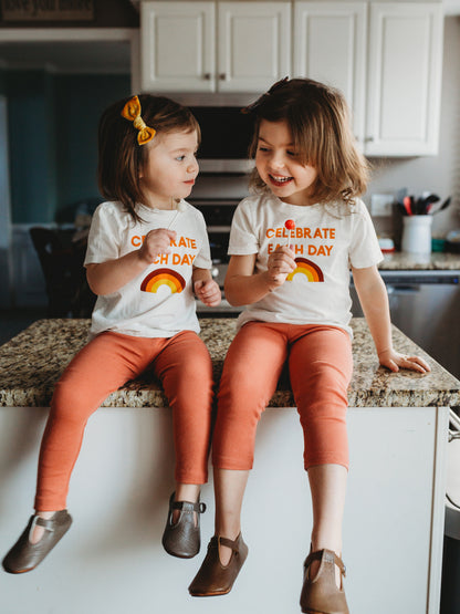 Two children seated together wearing matching Celebrate Each Day rainbow tees.