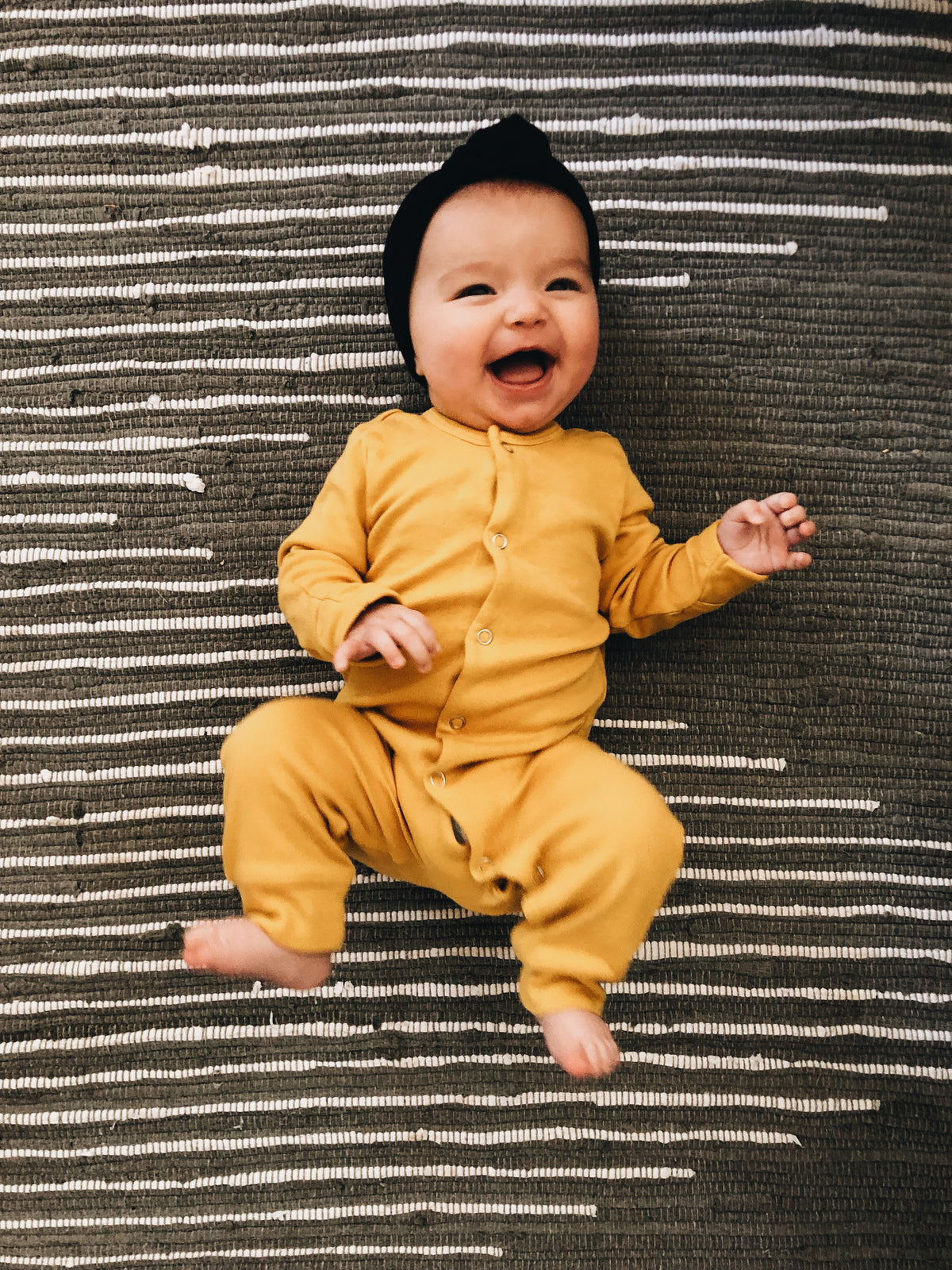 Full view of a smiling baby in a yellow coverall on a striped rug