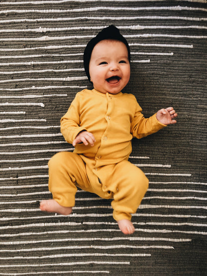 Full view of a smiling baby in a yellow coverall on a striped rug