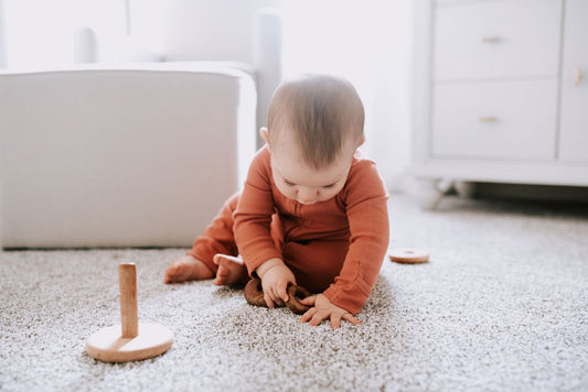 Baby in a rust-colored thermal zip coverall playing with wooden stacking rings on the floor.