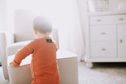 Back view of a baby standing beside a chair in a rust-colored thermal zip coverall.