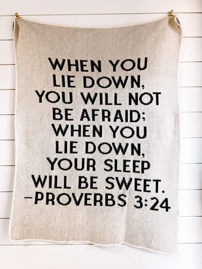 Natural Proverbs 3:24 blanket with black block lettering, hanging against a white plank wall.