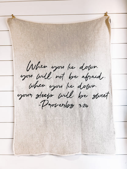 Natural Proverbs 3:24 blanket with black handwritten scripture, hanging against a white plank wall.