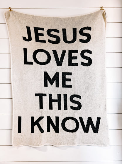 Natural Jesus Loves Me This I Know blanket with large black block lettering, hanging against a white plank wall.
